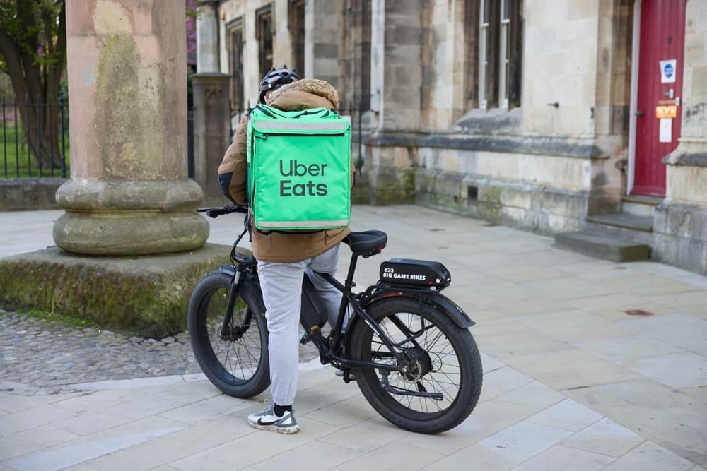 Uber Eats delivery cyclist with bright green bag, standing by a historic stone building.